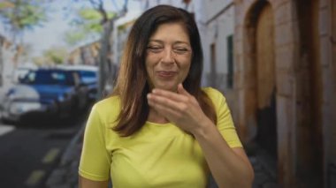 Kadın, klasik arabalar ve eski taş binalarla dolu güneşli bir caddede elleri ile ağzını kapatıyor; sessizlik..