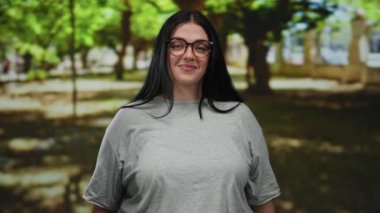 Parkta gülümseyen gözlüklü genç bir kadın. Güneşli yeşil ağaçlar ve gri bir tişörtü var. Kendine güveni ve sıcaklığı var..