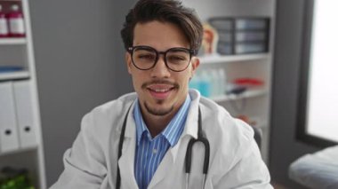 Gözlüklü ve steteskoplu genç adam klinik bir ortamda güvenle gülümsüyor, raflar ve tıbbi malzemelerle profesyonel bir tıbbi ortam sergiliyor..