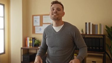 Kapalı alanda gülümseyen genç bir adam, raflarda kitap ve bitkiler bulunan bir ofis odasında sörfçü jestleri yaparak, pozitiflik ve günlük atmosfer aktarıyor.