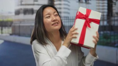 Şehir caddesinde kırmızı kurdeleli bir hediye kutusuyla gülümseyen bir kadın, açık hava ortamında genç Çinli bir kadınla neşeli bir an sergiliyor..