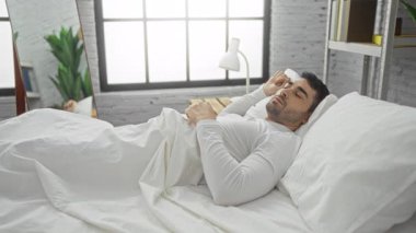 Genç bir adam, modern bir yatak odasında, bir yatakta oturuyor. Geniş bir pencereden gelen doğal ışıkla, rahat bir sabah rutini sergiliyor..