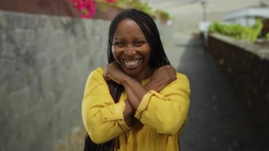 Kollarıyla gülümseyen kadın şehir sokağında sarı gömlekli, açık havada kıvırcık saçlı, gün içinde mutlu bir ifade sergiliyor..