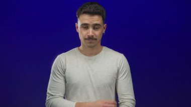 Genç İspanyol bir adam kollarını çapraz bir şekilde mavi bir arka plan duvarına yaslar, rahatlık ve iç gözlem yayar..