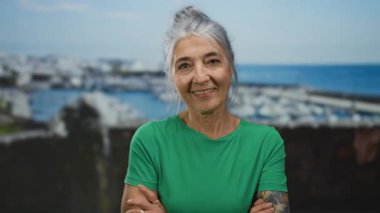 Gri saçlı, dövmeli ve yeşil gömlekli son sınıf öğrencisi kadın deniz kenarında kollarını kavuşturuyor, arkada açık mavi bir gökyüzünün altında tekne ve okyanusla gülümsüyor..