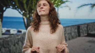 Güneşli bir sahil gezisinde gülümseyen ve el sallayan bir kadın canlı bir ifadeye sahip rahat bir kazak giyiyor ve okyanus arkaplanı neşeli bir açık hava anını yakalıyor..