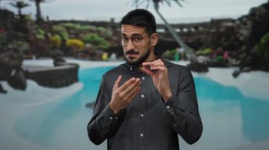 İspanyol bir adam lüks bir tatil köyündeki sakin bir havuzda, yemyeşil ve tropikal ambiyansla çevrili bir yerde el kol hareketi yapıyor..