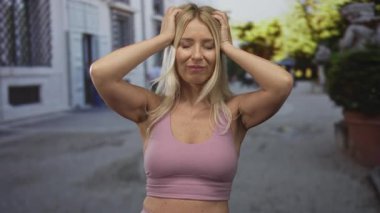 Pembe spor sutyen giyen sarışın kadın saçlarını tutuyor ve gündüz vakti binaların önünde ellerini çırılçıplak geziyor..