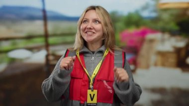 Kırmızı yelek giyen ve gönüllü rozet takan bir kadın şehirdeki hareketli bir açık hava restoranının terasında dikiliyor. Etrafı canlı çiçekler ve şehir manzarasıyla çevrili..