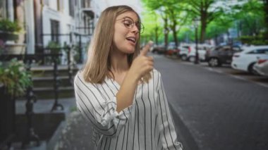 Genç sarışın kadın, çizgili bluzuyla ağaç kaplı bir caddede sağı işaret ediyor..
