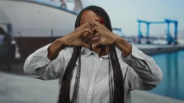 Limanda gülümseyen bir kadın elleriyle kalp hareketi yapıyor. Manzaralı tekne ve deniz manzarasına karşı sevgi ve mutluluk sergiliyor..