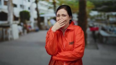 Turuncu hapishane tulumlu bir kadın, şehir manzarası arkasında bulanık bir şekilde, sıkıntılı görünen bir sokakta duruyor, bir anlık duygu ve hassasiyeti yakalıyor..