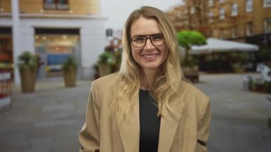 Sarışın bir kadın şehirde komik suratlar yapıyor, gözlük takıyor ve bej bir ceket giyiyor, arka planda şehir manzarası var, neşeli ve neşeli bir tavır sergiliyor..