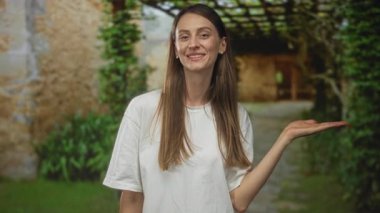 Avuç içiyle gülümseyen bir kadın bina girişinde açık elle sunar; dostça karşılama.