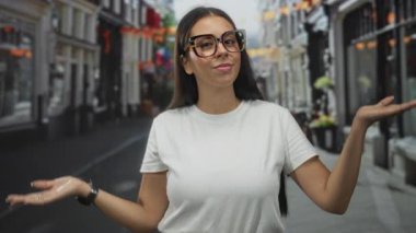 Büyük boy kaplumbağa kabuğu gözlüklü, beyaz tişörtlü genç esmer kadın avuçlarını kaldırdı ve sokakta omuz silkti..