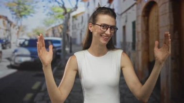 Elleri havada bir kadın sokakta park etmiş arabanın yanında gülümseyen gözlüklü ve kolsuz tişörtlü bir kadın..