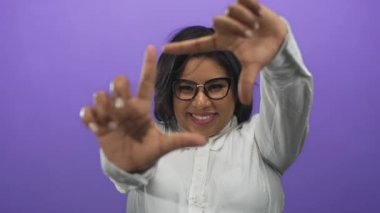 Elleriyle gülümseyen ve çerçeveleyen bir kadın, mor arka planda gözlük takan, izole bir ortamda pozitiflik ve yaratıcılık taşıyan bir kadın..