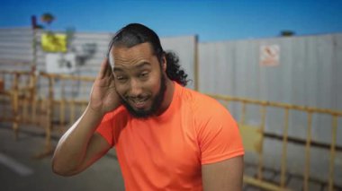 Parlak turuncu gömlekli bir adam inşaat bariyerinin önünde el ele tutuşuyor. Meraklı bir şekilde dinliyor..