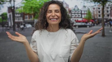 Kadın, şehir binalarının önünde, yeşil ağaçların altında, parlak bir gülümsemeyle el çırpıyor; mutluluk..