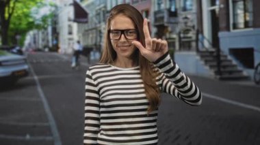 Kadın işaret parmağı tutuyor ve gözlüklü ve çizgili gömlekli bir caddede gülümsüyor. Kendine güvenen bir oyuncu..