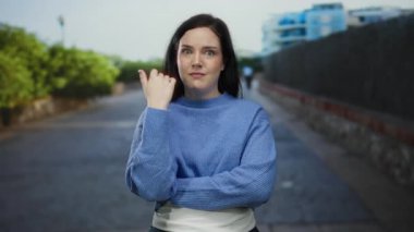 Esmer saçlı ve mavi kazaklı bir kadın, arka planda yeşil ağaçlar ve modern mimariyle şehir sokaklarında duygularını ifade ediyor..