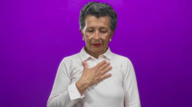 Beyaz tişörtlü, gri saçlı son sınıf öğrencisi bir kadın duygularını ifade ediyor. Göğsünde canlı mor bir arkaplan var. Gözleri yavaşça kapanıyor..