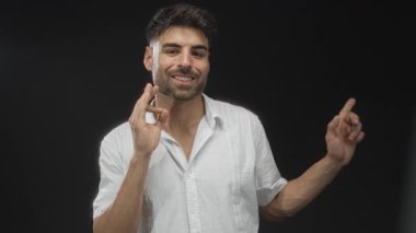 Man gesturing ok sign with right hand and pointing finger with left hand in studio against black backdrop; confidence approval.