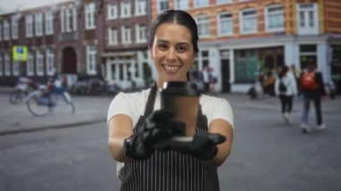 Önlüklü bir kadın bir kafe önünde kağıt bardak tutuyor, gülümsüyor ve içki ikram ediyor; arkadaşça bir servis..