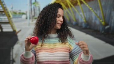 Kırmızı kalbi olan bir kadın renkli süveter ve kıvırcık saçlarıyla sokakta gülümsüyor, başparmağıyla işaret ediyor, açık şehir ortamında pozitifliğini ifade ediyor..
