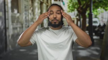 Bir şehir caddesinde elleri havada ve sıkılmış sakallı bir adam geniş bir gülümseme ve beyaz bir tişört giyen şehir dışında ileriye bakıyor; sevinç kutlaması enerjisi.