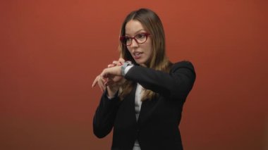Siyah ceketli genç bir kadın akıllı olanı kontrol ediyor ve turuncu stüdyoda çıplak elle bileği işaret ediyor; zaman baskısı sabırsızlığı.