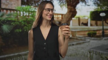 Bir kadın kahve fincanı tutuyor ve ağaçlarla dolu bir caddede çeşmenin yanında gülümsüyor..