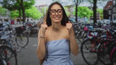 Çıplak omuzlu ve askısız elbisesi olan genç İspanyol kadın Amsterdam 'da bisikletlerin yanında baş parmağıyla gülümsüyor..