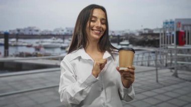 Sahildeki rıhtımın yakınındaki bir limanda elinde kağıt kahve fincanı tutan kadın, gözleri kapalı gülümsüyor ve fincanı sunuyor; keyif kahve molası..