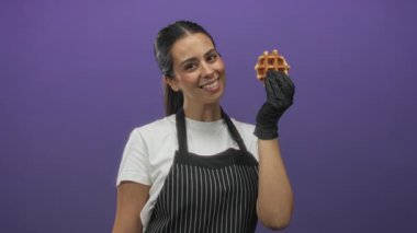 Önlüklü kadın, gülümseyen ve eldivenli bir elle waffle 'ı tutarken mor stüdyoda waffle' ı işaret ediyor..