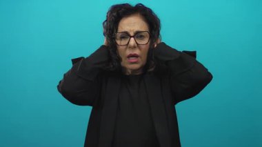 Orta yaşlı, gözlüklü İspanyol kadın yeşil arka planda kulaklarını kapatıyor. Canlı bir duvarın önünde dururken stres ya da hayal kırıklığını ifade ediyor..