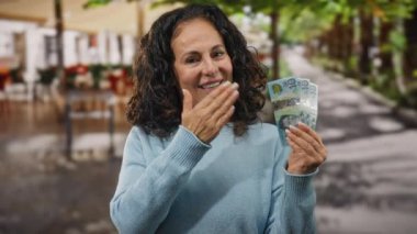 Bir şehir caddesinde Avustralya doları tutan, gülümseyen ve onay veren, pozitiflik ve finansal başarı taşıyan, güneşli bir günde orta yaşlı bir kadın..