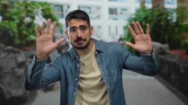 Kentsel alanda sakallı genç bir adam gün ışığında yeşil bir zemin ve binalara karşı şaşkınlığını dile getiriyor. Günlük kot ve bej giysiler giyiyor..