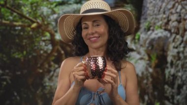 Orta yaşlı kadın bikini giyiyor ve güneş şapkası takıyor. Açık havada, yemyeşil park ortamında içmekten hoşlanıyor.