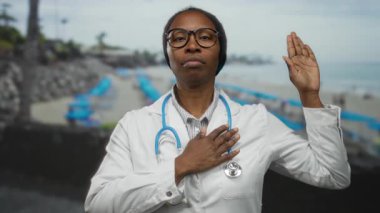 Steteskoplu kadın doktor plajda yemin ediyor. Huzurlu deniz manzarasındaki tıbbi bağlılığı simgeliyor..