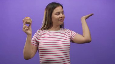 Keki sol eliyle tutan ve sağ eliyle mor stüdyoda çizgili tişört giyen neşeli bir kutlama ikramı sunan kadın..