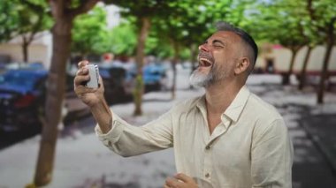 Akıllı telefonu elinde tutan adam sokakta ekrana bakarken gülüyor ve göz siliyor; neşe bağlantısı.