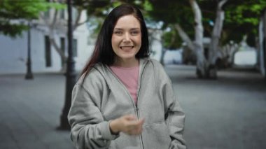 Esmer saçlı, gri kapüşonlu ve pembe tişörtlü, ağaçlar ve şehir manzarasıyla çevrili, açık hava yaşam tarzını gözler önüne seren eski şehrin sokaklarında gülümseyen bir kadın..