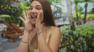 Güneşli bir parktaki yemyeşil ağaçların arasında haykırırken kız ellerini açık ağzının etrafına koyuyor; coşku.