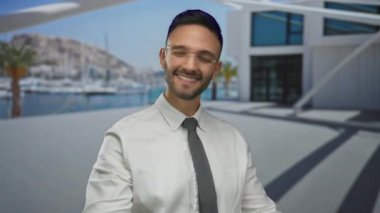 Genç adam deniz kenarında neşeyle gülümsüyor, gözlük takıyor ve modern mimari geçmişi ve marinası olan bir kravat takıyor..