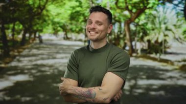 Genç adam açık hava parkında gülümsüyor ağaçlar ve güneş ışığı dallardan süzülüyor, dövmeli kolunu ve günlük tarzını gösteriyor..