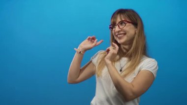 Kırmızı gözlüklü, beyaz tişörtlü ve mavi stüdyoda gülümseyen bir kadın..