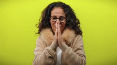 Parlak sarı arka planda dua eden bir kadın gözlüklü rahat bir palto giyiyor, kıvırcık saçları umut, sükunet ve içgözlem ifadesini çerçeveliyor..