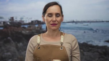 Önlük takan kadın, açık bir gökyüzünün altında canlı bir okyanus zeminine kurulmuş çeşitli duyguları ifade eden kayalık bir sahil kenarında duruyor..