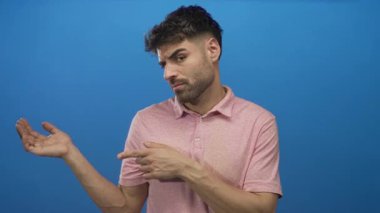 Young hispanic man in pink polo shirt points finger to palm showing hand gesture in blue studio setting; doubt.